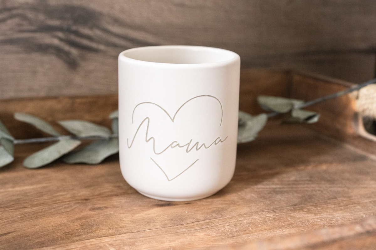 Matte weiße Keramiktasse mit Herz-Gravur und personalisiertem Schriftzug „Mama“ auf Holzuntergrund, Eukalyptus im Hintergrund.